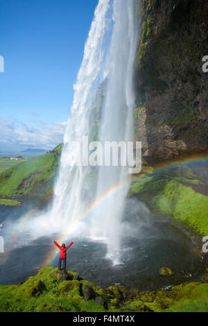 Person and rainbow beneath 60m-high Seljalandsfoss waterfall, Sudhurland, Iceland. Stock Photo