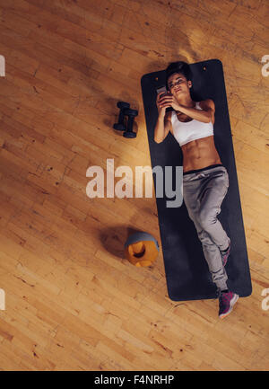 Overhead view of fitness woman lying on mat using mobile phone. Top view of young female taking break from exercise. Stock Photo