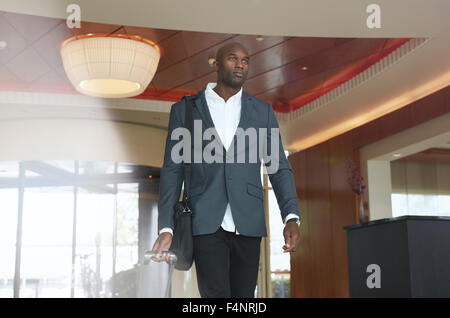 Portrait of young businessman with luggage in hotel lobby. African business executive walking in hotel hallway with his baggage. Stock Photo