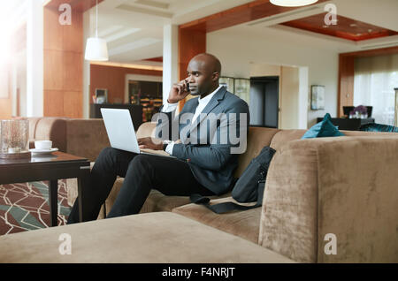 Businessman sitting on sofa with a laptop using mobile phone. Busy african male executive waiting in hotel lobby. Stock Photo