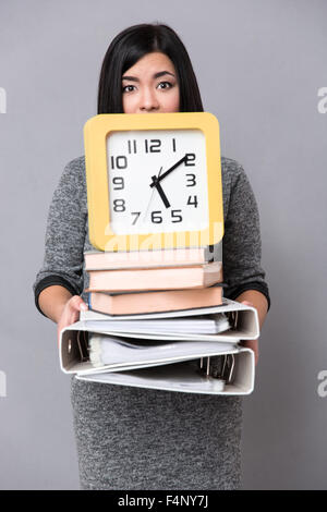 Portrait of a young woman holding books, folders and wall clock over gray background Stock Photo