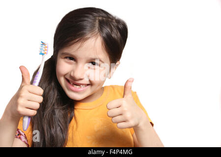 Little seven year old girl shows big smile showing missing top front teeth and holding a toothbrush with thumbs up on a white ba Stock Photo