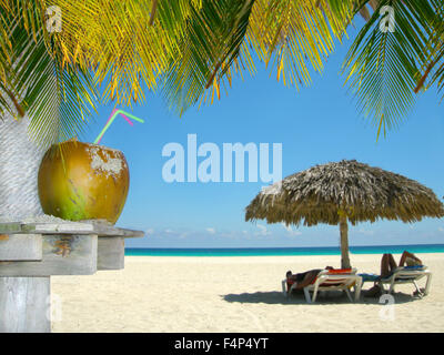 People relaxing under tropical huts with coconut and palm leaves in the foreground in a Cuban beach Stock Photo