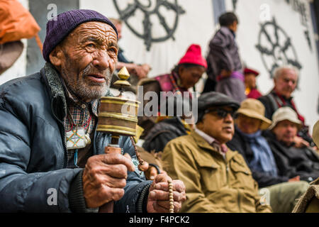 An old ladakhi man is turning a prayer wheel in the courtyard of Hemis Gompa, watching monks with big wooden masks and colorful Stock Photo