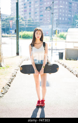 Young handsome asiatic long brown straight hair woman skater posing outside in the city, looking in camera holding a skateboard, serious - pensive, thoughtful, thinking future concept Stock Photo
