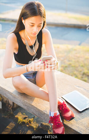 Young handsome asiatic long hair woman sitting on small wall, listening music with earphones, using smartphone looking down the Stock Photo