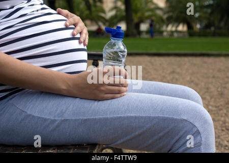 Pregnant woman outdoors holding bottle of water. She is sitting in the park Stock Photo