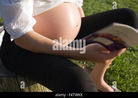 High view of a young pregnant woman sitting on a trunk close to grass and reading book Stock Photo