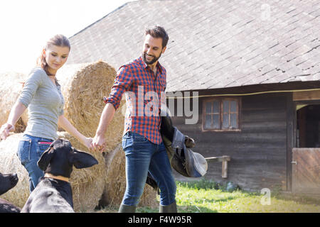 Couple holding hands walking with saddle and dogs outside barn Stock Photo