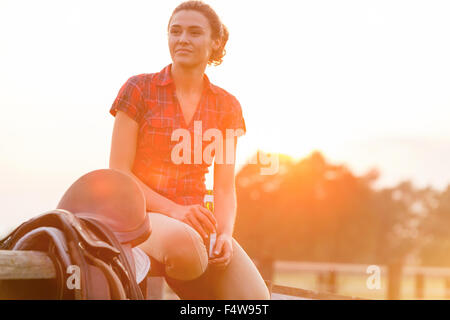 Smiling woman drinking beer next to saddle on rural fence Stock Photo