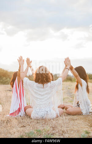 Boho women sitting in circle with arms raised and connected in rural field Stock Photo