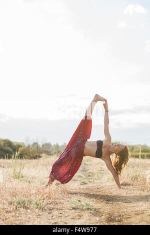Boho woman in extended side plank yoga pose in sunny rural field Stock Photo