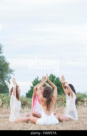 Boho women meditating with hands clasped overhead in circle in rural field Stock Photo