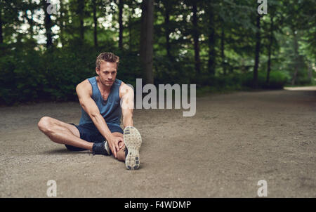 Handsome young male athlete limbering up before training sitting on the ground in a wooded park doing stretching exercises in a Stock Photo