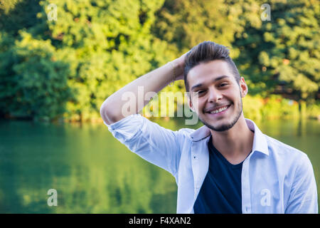 Portrait of contemplative light brown haired young man wearing white shirt beside picturesque river or lake, smiling to camera Stock Photo