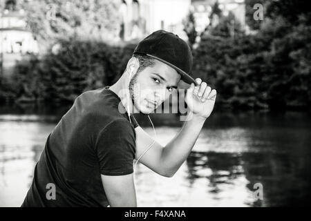 Portrait of contemplative light brown haired young man wearing baeball cap, beside picturesque river or lake, looking at camera Stock Photo