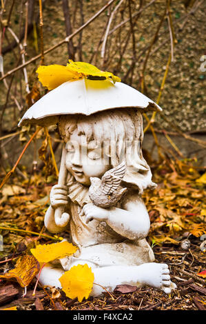 Profile of a small weathered statue of a young girl holding an umbrella and a bird, surrounded by dry leaves and bark chips. Stock Photo