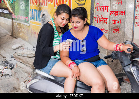 Two young indian women in shorts are sitting on a scooter and using a mobile phone Stock Photo