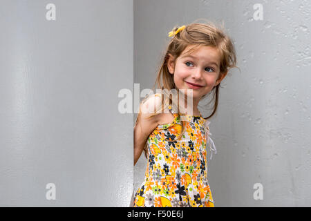 A smiling  three year old girl, wearing a yellow dress, is standing between silverish walls Stock Photo