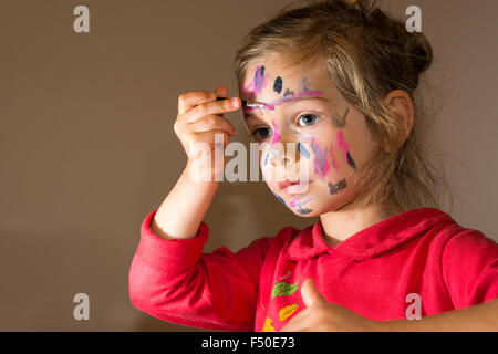 A little girl, wearing a red sweater, is painting her face with colorful watercolor Stock Photo