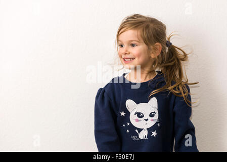 A Portrait of a smiling three year old girl with fair hair, wearing a blue sweater with the picture of a white cat Stock Photo