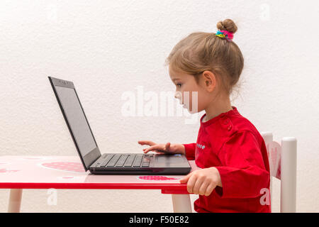 A blond three year old girl is sitting in front of a notebook, laptop, watching the screen and using the keyboard Stock Photo