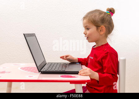 A blond three year old girl is sitting in front of a notebook, laptop, watching the screen and using the keyboard Stock Photo