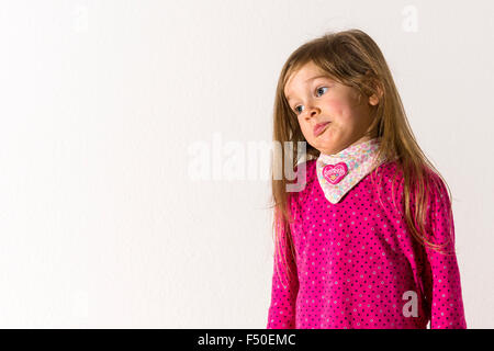 A blond three year old girl, wearing a pink shirt, is looking skeptical in front of a white wall Stock Photo