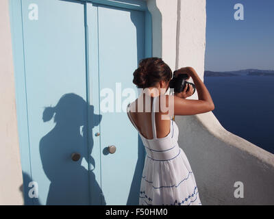 A young woman in a white dress takes pictures of the Aegean Sea from a building in Santorini, Greece Stock Photo