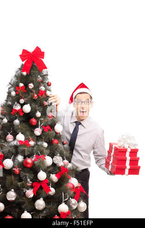 Vertical shot of a joyful man holding three Christmas presents and hiding behind a Christmas tree isolated on white background Stock Photo