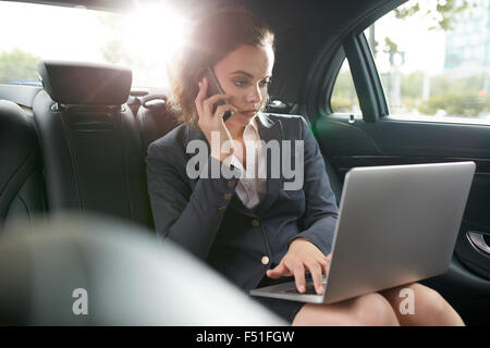 Young businesswoman talking on cell phone sitting in back seat of car. Female CEO using laptop and answering phone call while tr