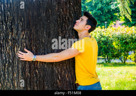 Smiling handsome young man hugging a tree, looking up, in city park Stock Photo