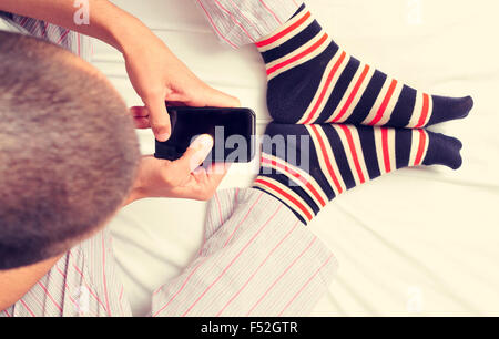high-angle shot of a young caucasian man wearing pajamas and colorful striped socks using a smartphone in bed Stock Photo