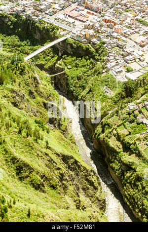 An aerial photograph captures a deep, narrow river canyon flanked by steep, green-covered cliffs. A concrete bridge spans the canyon, connecting a densely built town on the upper edge to the opposite Stock Photo