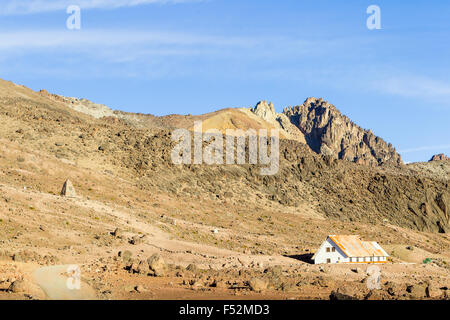 A small white cabin with a rusted metal roof sits in a barren, rocky desert valley beneath rugged, jagged mountains. The terrain is dry and sparsely vegetated, with scattered boulders and a dirt path Stock Photo