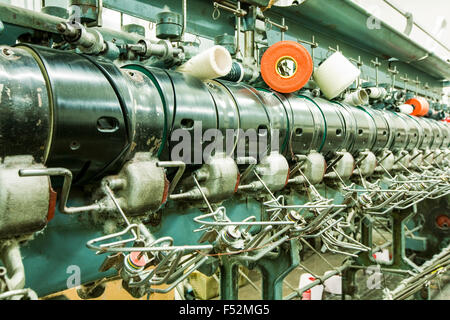 A close-up view of a large industrial textile spinning machine in operation, featuring a row of metallic spindles with white and orange thread spools mounted on top. Thin threads are being drawn from Stock Photo
