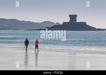 Pensioner couple walking along the shore line on St St Ouen's beach with La Rocco Tower in the background Jersey Channel Islands Stock Photo