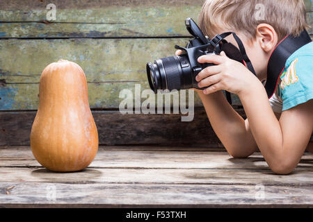 Funny fair boy use DSLR camera trying to take photo of ripe pumpkin on wooden background in studio. Photography lessons or learn Stock Photo