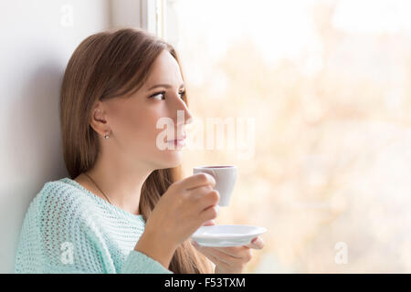 Young woman drinking coffee near the window on sunny day. Pretty adult thinking girl holds cup of espresso and looking through w Stock Photo