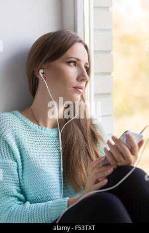 Young woman listening to music in her smartphone and looking through window. Beautiful girl relax at window sill. Stock Photo