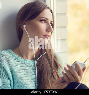 Young smiling woman having fun with listening to music or using her smartphone. Happy beautiful girl relax at window sill. Warm Stock Photo