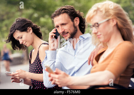 A man and two women seated on a bench in a park, checking their phones, one making a call. Stock Photo