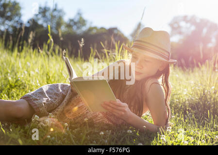 A young girl in a straw hat lying on the grass reading a book. Stock Photo