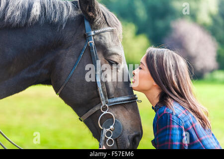 Beautiful happy Asian Eurasian young woman or girl wearing plaid checked shirt, smiling and resting her head on her horse in sun Stock Photo