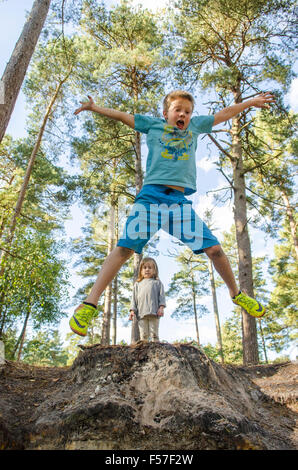 nine year old boy doing star jumps off a bank with his three years old sister watching in woodland, Sussex. UK. October. Stock Photo