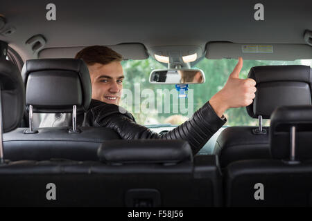 Happy young man behind the steering wheel looking towards the back seat and giving a thumbs up. Stock Photo