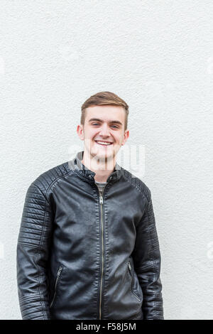 Medium shot of a young man wearing a leather jacket standing behind a white wall. Stock Photo