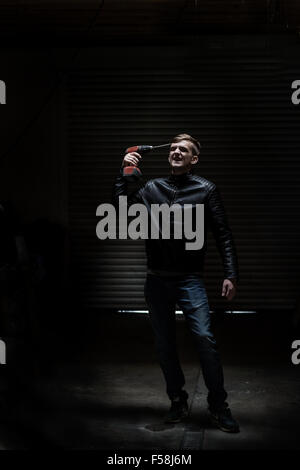 A young man points an electric screwdriver at his temple while standing behind a metal door in a garage. Long shot. Stock Photo