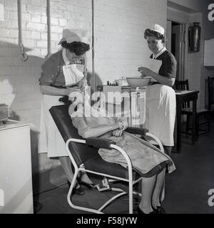 1950s, historical picture, a young female factory worker in her overalls sitting back in a chair having medical attention, her mouth and teeth are being examined by the work's matron, with a nurse in attendance. Stock Photo