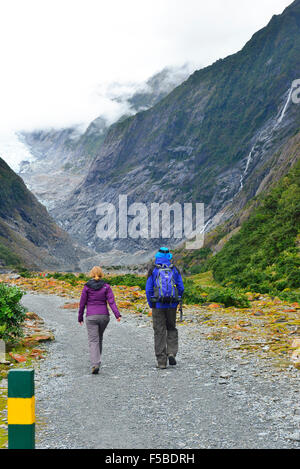 Two hikers walking along  a rocky riverbed path next to  glacier melt water river up to the Toe of the Franz Josef glacier South Island, New Zealand Stock Photo